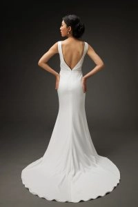 A young woman wearing a sleek, white wedding dress with a deep V-back and a fitted silhouette that flows into a small train. She stands with her hands on her hips, showcasing the elegant lines of the dress from behind. Her hair is styled in an updo, and she wears delicate earrings. The dark background accentuates the dress's sophisticated design and her graceful posture.