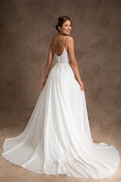 A young woman is wearing a beautiful white wedding Lacy dress with a lace bodice and thin straps. The back of the dress features a deep V-cut, button details, and a flowing skirt that creates an elegant train. She has her hair styled up with a decorative hairpiece and is smiling while looking back over her shoulder. The background is a plain brown, highlighting her and the dress.