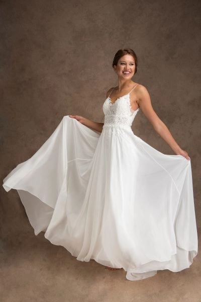 A young woman wearing a beaded chiffon wedding dress with a lace bodice and thin straps. She holds the sides of the flowing skirt, smiling brightly. Her hair is styled up with a decorative hairpiece, and the plain brown background accentuates the elegance of the dress and her joyful expression.