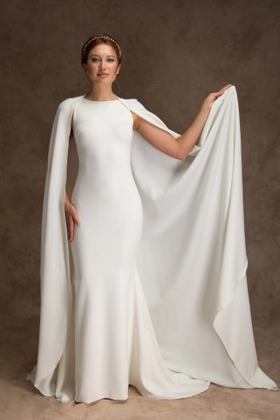 A woman poses in a sleek white bridal gown with a high neckline and flowing train. She wears a dramatic bridal cape and a pearl-accented headband, adding a regal touch to her elegant look.