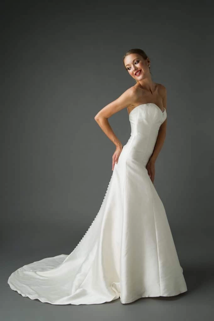 A young woman wearing a strapless trumpet wedding dress that elegantly hugs her figure and flares out at the bottom. The dress features a smooth, satin finish with a row of buttons running down the back. She poses with a smile, hands on her hips, and her hair styled in an elegant updo. The dark background highlights the sleek lines and sophisticated design of the dress.