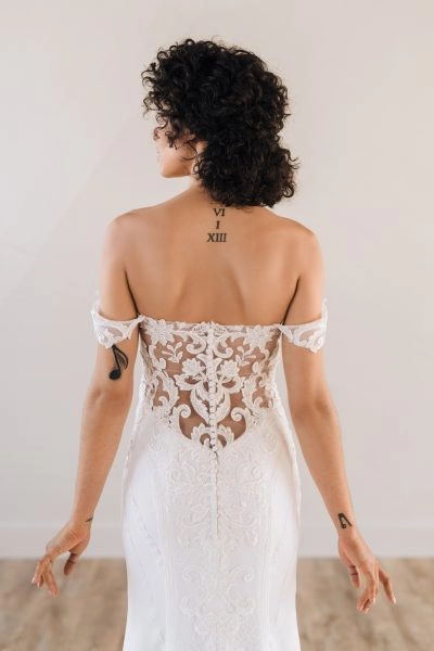 A woman wearing a lace off-the-shoulder wedding dress. The dress features intricate lace detailing along the back, with delicate buttons running down the center. The off-the-shoulder sleeves add a romantic and elegant touch, complementing the fitted silhouette of the gown. The lace pattern is beautifully designed to highlight the curves, and the sheer elements add sophistication to the overall look.