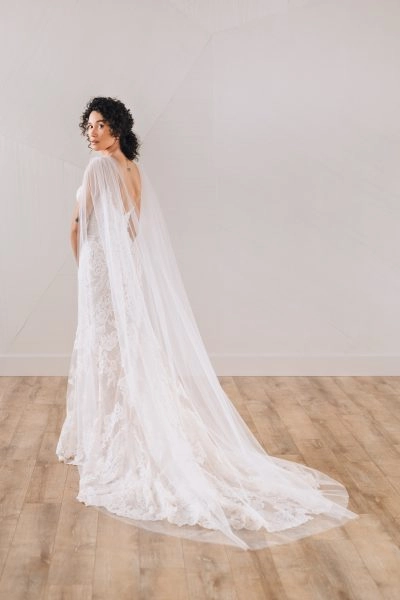 A bride in a lace gown wears a Tulle Wedding Cape with a raw edge, pinned with delicate gold pearl pins. She stands on a wooden floor, looking over her shoulder.