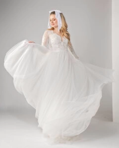 Flowing long sleeve lace and tulle wedding dress with a lace bodice and soft, airy skirt. The bride gracefully lifts the skirt, showing off the dress's movement, while a large white bow in her hair adds a charming, vintage-inspired accent to her look.
