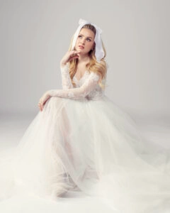 Whimsical long sleeve lace and tulle wedding dress, featuring a delicate lace bodice and flowing tulle skirt. The bride sits thoughtfully, wearing a large white bow in her hair, adding a playful and vintage-inspired touch to her bridal look.