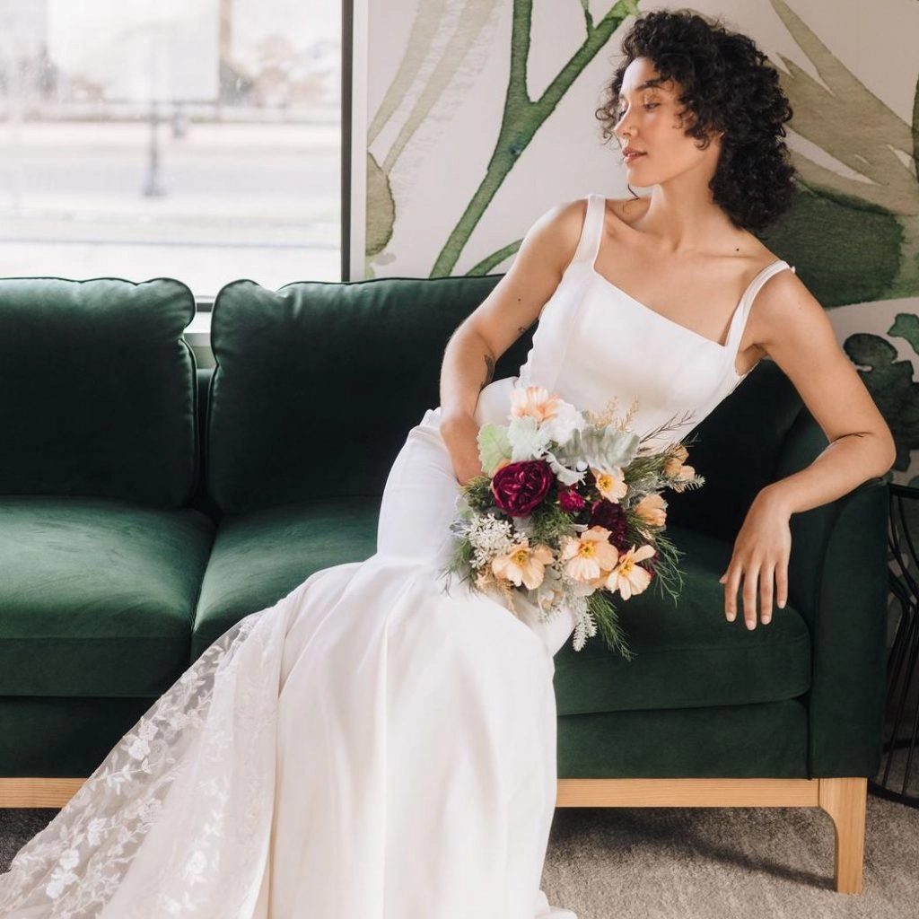 Bride wearing a square neck trumpet wedding dress holding a bouquet with peach flowers sitting on a green couch
