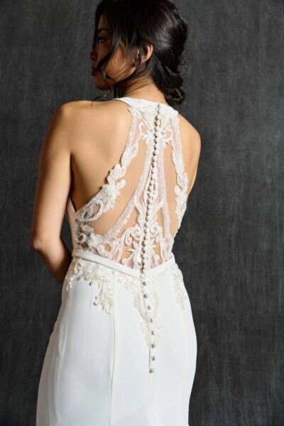 A woman wearing a beaded back wedding dress. The back of the dress features intricate lace and beadwork, with sheer panels and a delicate row of buttons running down the center. The beaded details form elegant patterns along the edges of the dress's open back, adding a sophisticated and elegant touch to the design.