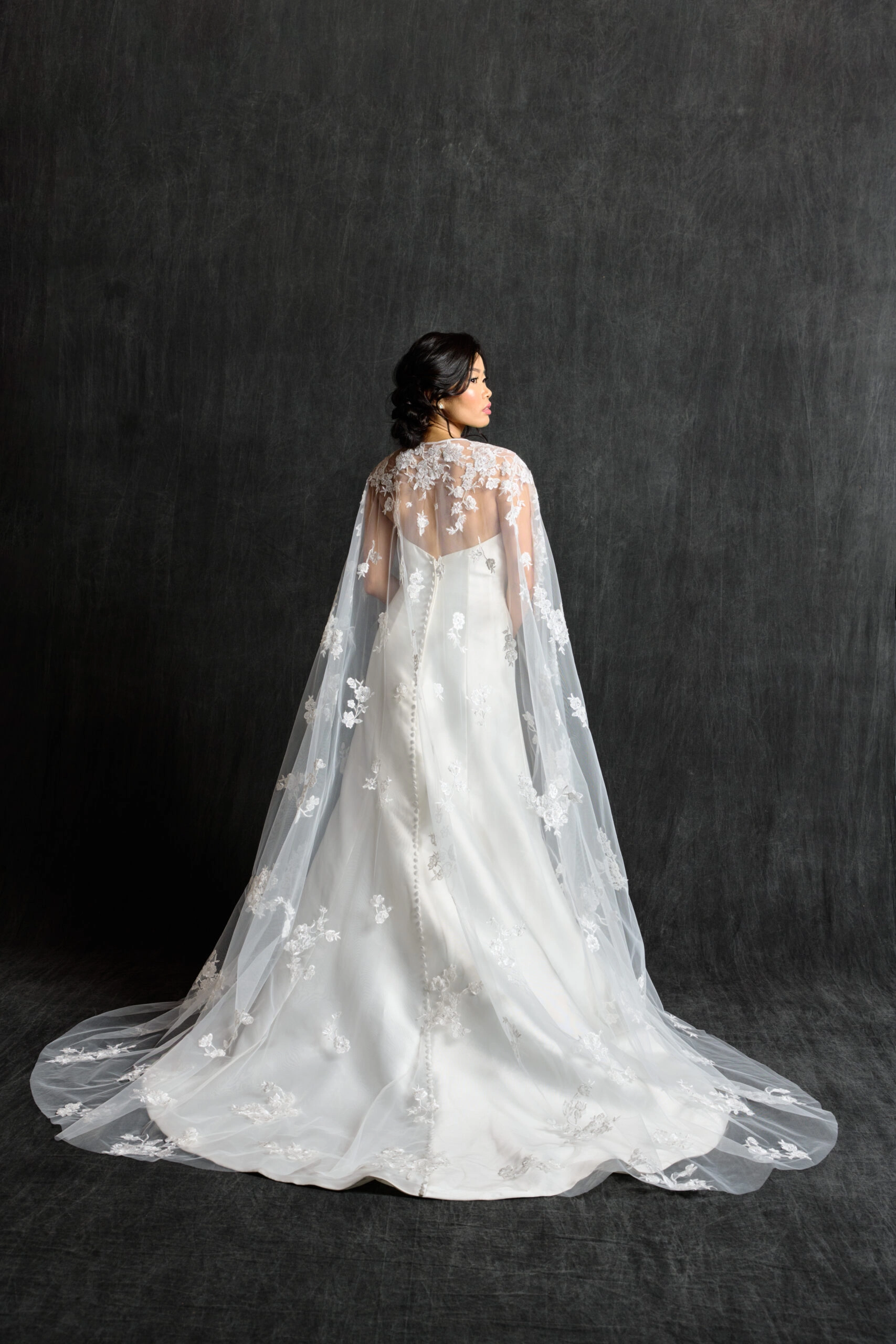 A bride is photographed from behind, showcasing the back of her fitted satin gown with a row of buttons running down the train. Draped elegantly over her shoulders is a sheer lace cape, embroidered with delicate floral details, adding a romantic and ethereal touch to the look.