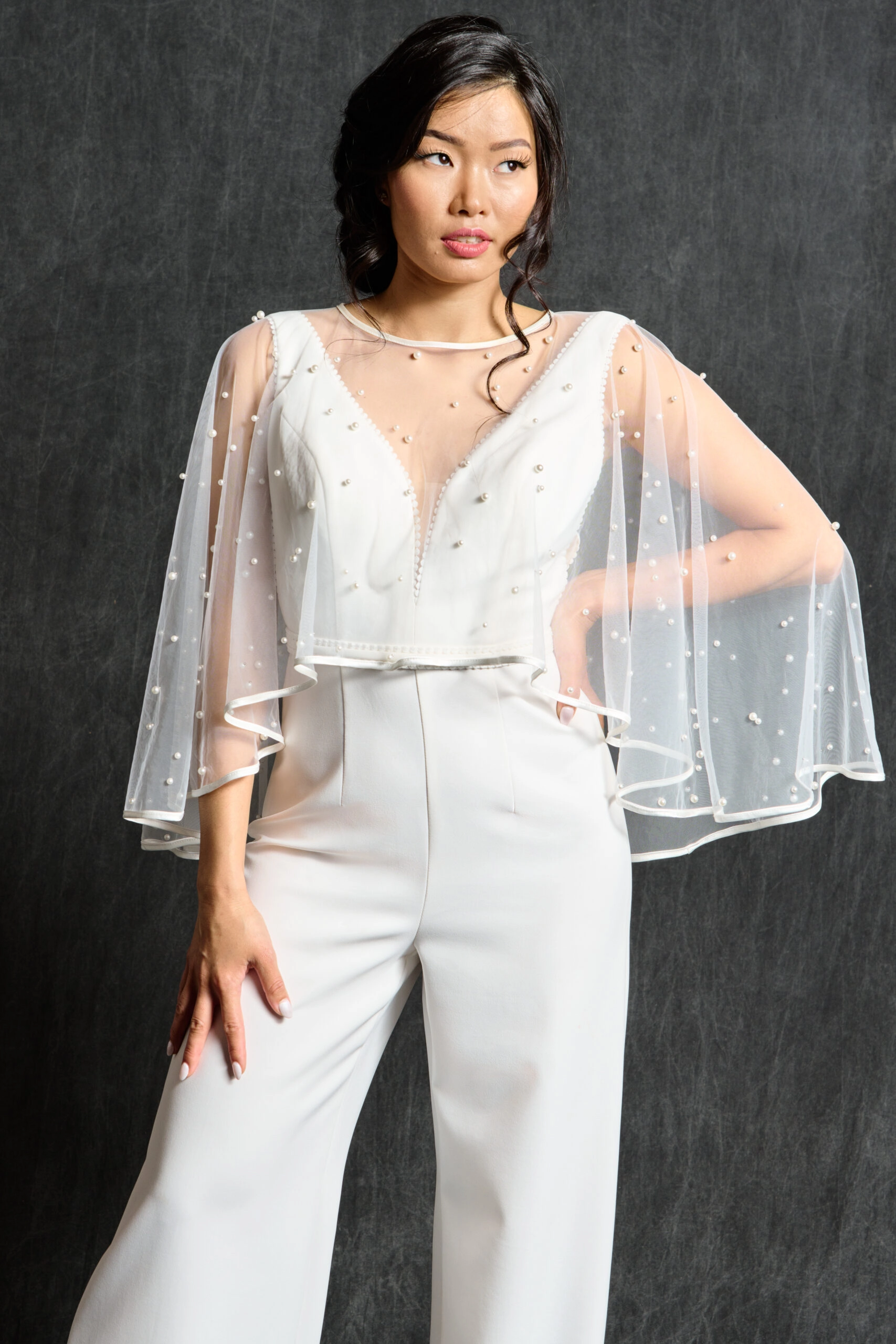 A bride is shown wearing a bridal jumpsuit paired with a delicate beaded capelet. The capelet, made from sheer fabric, is adorned with pearls, adding a soft, romantic touch to the modern, structured outfit. The combination of the sleek jumpsuit and the detailed capelet creates a unique and contemporary bridal look.