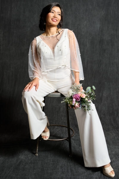 A bride is seated on a stool, wearing a chic, tailored bridal jumpsuit paired with an elegant beaded bridal capelet. The sheer capelet is embellished with pearls, adding a touch of modern sophistication to her look. She holds a small, colorful bouquet in her hand, completing her stylish and contemporary bridal ensemble.
