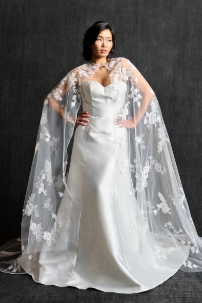 A model stands confidently in a sleek satin wedding gown, complemented by a sheer lace cape adorned with floral appliqué. The cape adds a romantic and regal touch to the clean lines of the dress, creating an elegant and modern bridal look.