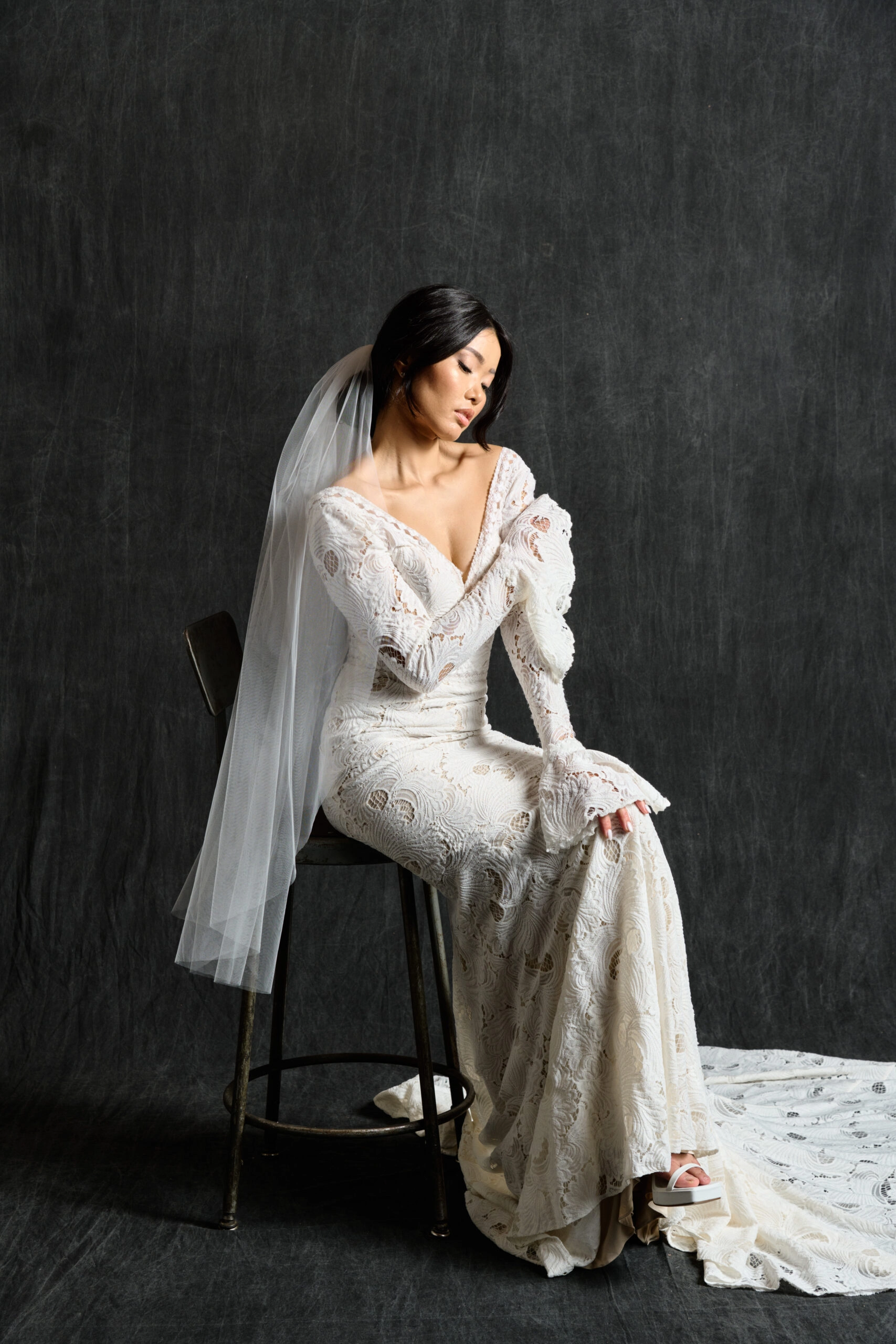 A bride sits gracefully on a stool, wearing an intricately designed lace wedding gown with long sleeves and a deep V-neckline. The dress features a vintage-inspired pattern, adding a touch of elegance and sophistication. Her sheer veil falls gently over her shoulders, complementing the dress's detailed lacework. The backdrop is a dark, textured surface that highlights the delicate lace and the flowing train of the gown. The bride's pose and serene expression enhance the timeless beauty of the ensemble.