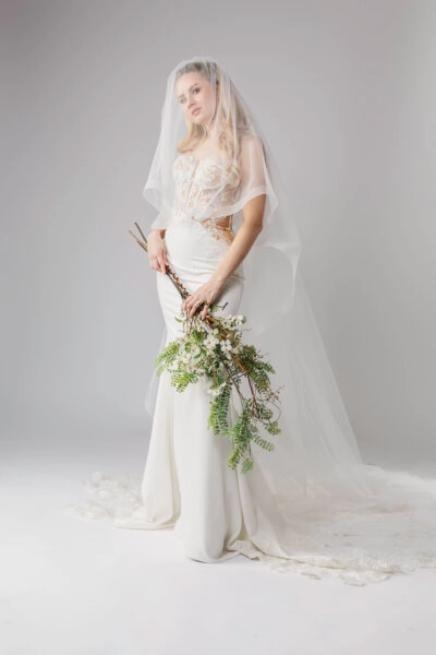 A lace corset wedding dress with a long veil. The dress features a structured lace bodice with visible corset boning and a fitted, smooth skirt that extends into a lace train. The woman holds a bouquet of wild branches and greenery, adding a natural, rustic touch to her elegant look. The soft, sheer veil frames her face, enhancing the romantic feel of the ensemble.