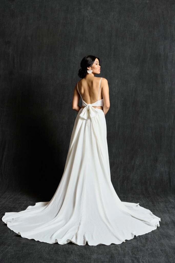 A contemporary bridal gown featuring delicate spaghetti straps and a deep V-back. The dress boasts a fitted bodice that transitions into a flowing, elegant skirt with a long train. The back is accentuated with a stylish bow, adding a touch of charm and sophistication. The minimalist design highlights the bride's graceful silhouette. The bride completes her look with chic earrings and a refined updo.