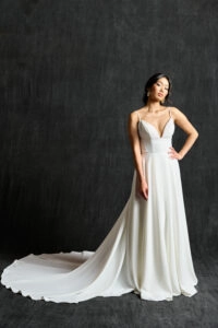 A modern bridal gown featuring delicate spaghetti straps and a plunging V-neckline. The dress has a fitted bodice that flows into a voluminous, elegant skirt with a long train. The clean, minimalist design highlights the bride's natural beauty and grace. The bride completes the look with simple, chic earrings, and her hair is styled in a sophisticated updo.