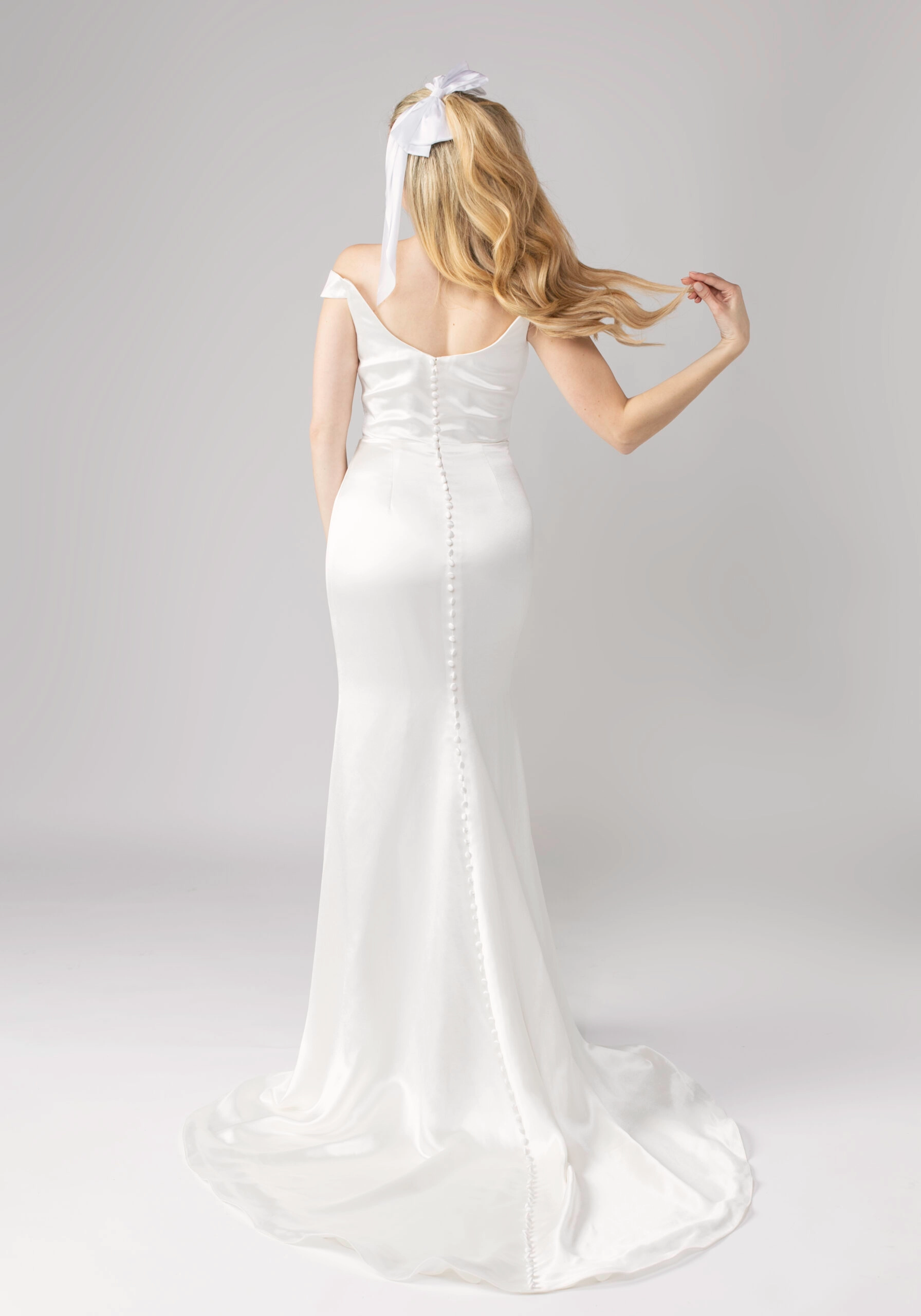 Full back view of satin off-the-shoulder wedding dress with button detailing and long flowing train.