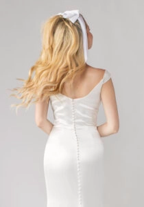 Back view of a satin off-the-shoulder wedding dress with buttons running down the train and cascading blonde curls tied in a white bow.