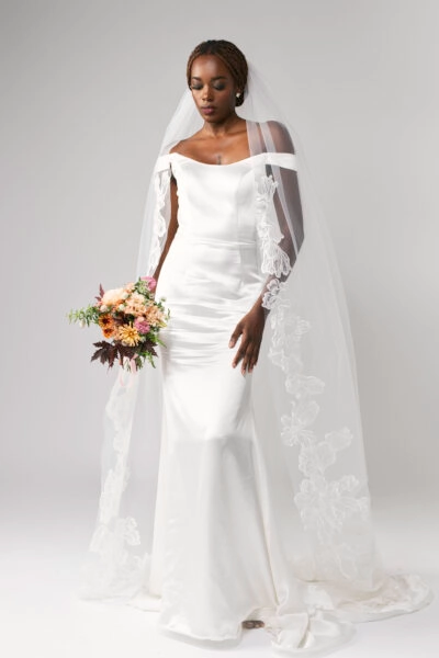 Woman in an off-shoulder wedding dress holding a bouquet, wearing a chapel length lace cathedral veil.