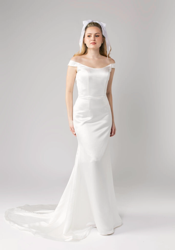 Elegant satin off-the-shoulder wedding dress with a fitted bodice and flowing train, modeled by a bride with a bow headband.