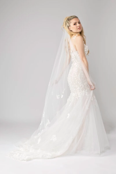 Woman in a lace wedding dress with a long 3D floral veil, looking over her shoulder.